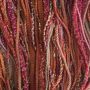 Puede incluir: Primer plano de una variedad de hilos y cordones de colores, incluyendo rojo, marrón, dorado y verde. Los hilos están dispuestos en un patrón aleatorio, creando un fondo texturizado y visualmente atractivo.