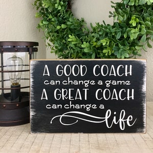 May include: A black wooden sign with white text that reads "A good coach can change a game. A great coach can change a life."