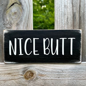 May include: Black wooden sign with white text that reads "NICE BUTT".