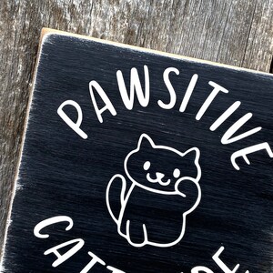 May include: A black wooden sign with white text and a cartoon cat illustration. The sign reads "PAWSITIVE CATITUDE" in a playful font, perfect for cat lovers. The sign has a distressed, rustic look.