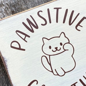 May include: A square, distressed white wooden sign with the words "PAWSITIVE ATTITUDE" in brown lettering. A cartoon cat illustration is in the center, waving its paw. The sign has a rustic, farmhouse aesthetic.
