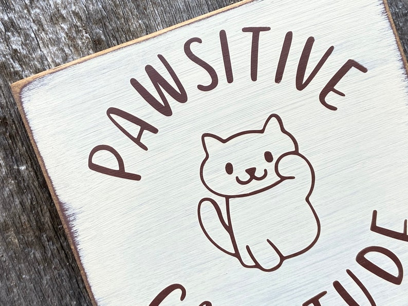 May include: A square, distressed white wooden sign with the words "PAWSITIVE ATTITUDE" in brown lettering. A cartoon cat illustration is in the center, waving its paw. The sign has a rustic, farmhouse aesthetic.