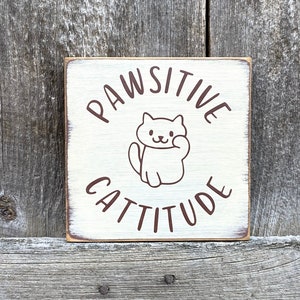 May include: A square, cream-coloured wooden sign with the words "PAWSITIVE CATTITUDE" in brown lettering. A cartoon cat illustration is centered on the sign. The sign has a distressed, rustic appearance, with visible wood grain.