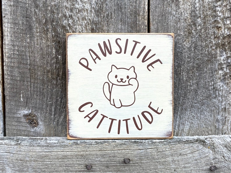 May include: A square, cream-coloured wooden sign with the words "PAWSITIVE CATTITUDE" in brown lettering. A cartoon cat illustration is centered on the sign. The sign has a distressed, rustic appearance, with visible wood grain.