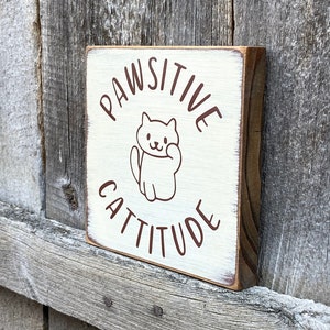 May include: A small, square wooden sign with the words "PAWSITIVE CATTITUDE" in brown lettering. A cartoon cat illustration is in the center. The sign has a distressed, white-painted surface and a natural wood edge.
