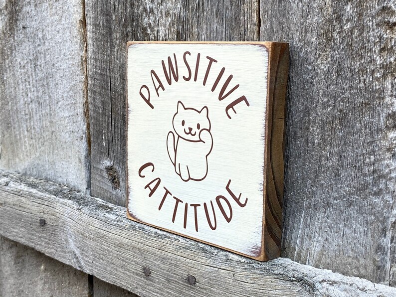 May include: A small, square wooden sign with the words "PAWSITIVE CATTITUDE" in brown lettering. A cartoon cat illustration is in the center. The sign has a distressed, white-painted surface and a natural wood edge.