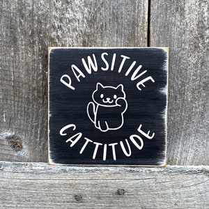 May include: Square black wooden sign with white text and a cat illustration. The sign reads "PAWSITIVE CATTITUDE" in a playful font. The cat illustration is a simple outline. The sign has a distressed, rustic appearance.