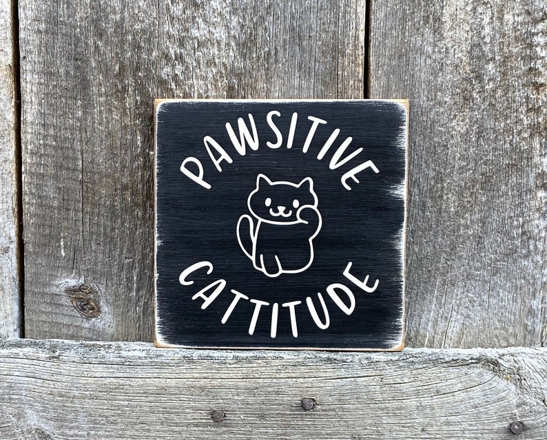 May include: Square black wooden sign with white text and a cat illustration. The sign reads "PAWSITIVE CATTITUDE" in a playful font. The cat illustration is a simple outline. The sign has a distressed, rustic appearance.