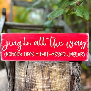May include: Red wooden sign with white lettering that reads "jingle all the way (NOBODY LIKES A HALF-ASSED JINGLER)"