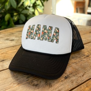 May include: A black and white trucker hat with the word "MAMA" in a floral pattern across the front. The hat has a black brim and a mesh back.