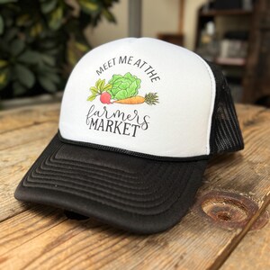 May include: A black and white trucker hat with the text "Meet Me at the Farmers Market." The hat features a white front panel with a colorful illustration of vegetables, including a carrot, radish, and lettuce.