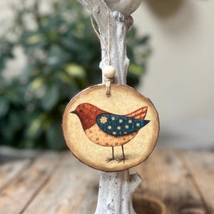 May include: A white bird figurine perched on a distressed white tree branch, with a round wooden ornament hanging below. The ornament features a colorful bird design with red, blue, and tan hues. The base is a distressed white pedestal.