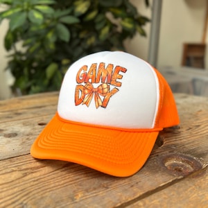 May include: A white and orange trucker hat with the words "GAME DAY" printed on the front. The text is in orange and features a basketball-themed bow. The hat has a curved brim and a mesh back.