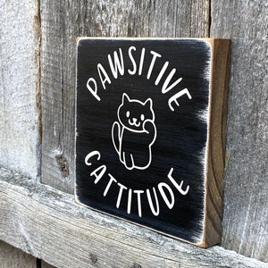 May include: A small, square wooden sign with a black painted surface. White text reads "PAWSITIVE CATTITUDE" with a cat illustration. The sign has a distressed, rustic look, with visible wood grain and a white border.