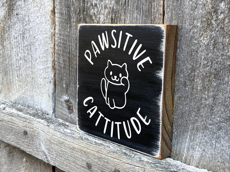 May include: A small, square wooden sign with a black painted surface. White text reads "PAWSITIVE CATTITUDE" with a cat illustration. The sign has a distressed, rustic look, with visible wood grain and a white border.