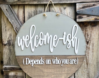 May include: Round wooden sign with the text "welcome-ish" in white script, with the phrase "(Depends on who you are)" below. The top half is painted gray, the bottom half is stained brown, and it hangs from a jute rope.