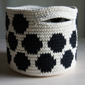 May include: A white crocheted basket with a black polka dot pattern. The basket has a handle and is made of thick yarn.