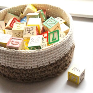 K&ouml;nnte beinhalten: Ein wei&szlig;er und brauner geh&auml;kelter Korb gef&uuml;llt mit bunten Holzw&uuml;rfeln mit Buchstaben und Bildern. Zwei W&uuml;rfel liegen au&szlig;erhalb des Korbs.