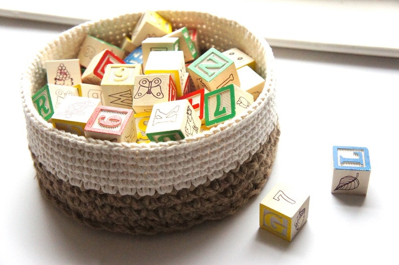K&ouml;nnte beinhalten: Ein wei&szlig;er und brauner geh&auml;kelter Korb gef&uuml;llt mit bunten Holzw&uuml;rfeln mit Buchstaben und Bildern. Zwei W&uuml;rfel liegen au&szlig;erhalb des Korbs.