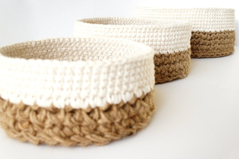 K&ouml;nnte beinhalten: Drei runde geflochtene K&ouml;rbe mit einem nat&uuml;rlichen Jute-Boden und einem wei&szlig;en geh&auml;kelten Oberteil. Die K&ouml;rbe sind ineinander verschachtelt, wobei der gr&ouml;&szlig;te Korb hinten und der kleinste Korb vorne ist.