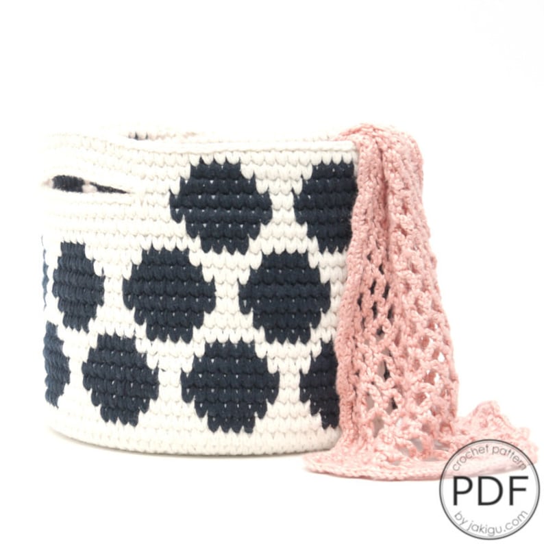 May include: A white crocheted storage basket with a black polka dot pattern. The basket is round and has a handle on the side. A pink crocheted bag is draped over the side of the basket.