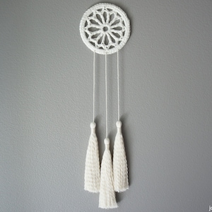 May include: White crocheted dream catcher with three white tassels hanging from it.