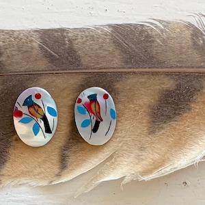 May include: A pair of oval-shaped earrings with a bird design. Each earring features a colorful bird perched on a branch with red berries and blue leaves, set against a white background. The earrings are displayed on a large feather.