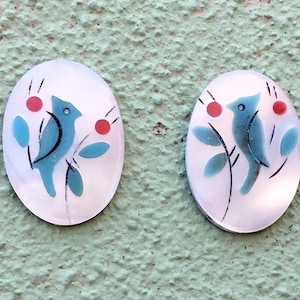 May include: A pair of oval-shaped ceramic earrings with a white background and a blue bird design. The bird is perched on a branch with red berries.