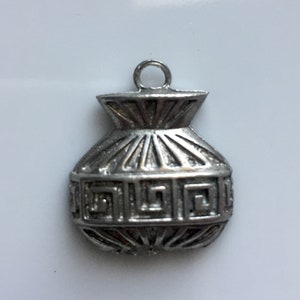 May include: A silver-colored charm in the shape of a small vase. The vase has a decorative geometric pattern around the middle and vertical lines radiating from the top. A small loop is at the top for attaching to a chain or bracelet.