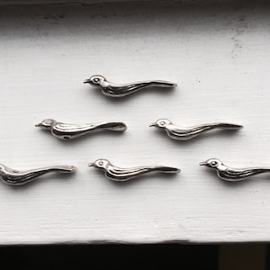 May include: Six small, silver-colored bird-shaped decorative embellishments are arranged on a white surface. The birds have a streamlined design with visible details like beaks and wings, suggesting a vintage or handcrafted style.