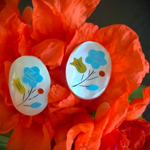 May include: A pair of white oval-shaped earrings with a blue, yellow and red floral design. The earrings are set against a backdrop of orange flower petals.