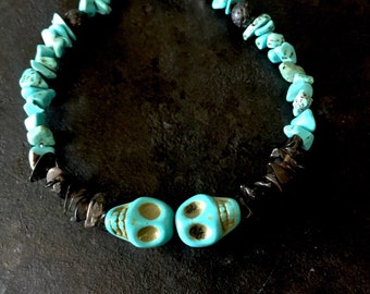 May include: A bracelet featuring two turquoise-colored skull beads, interspersed with irregular turquoise-colored stone chips, dark grey stone fragments, and a single black porous bead. The piece is secured with a silver-toned metal toggle clasp.