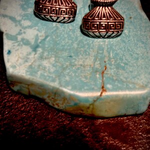 May include: Two silver-toned earrings with a vase-like design, featuring geometric patterns. The earrings are displayed on a turquoise stone, set against a dark brown background. The earrings have a vintage or tribal aesthetic.