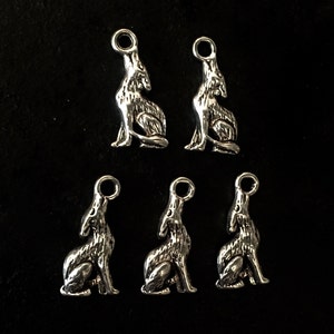 Op de afbeelding: Vijf zilverkleurige wolf bedels. Elke bedel toont een huilende wolf in een zittende positie, met een lus aan de bovenkant om aan sieraden of andere knutselwerken te bevestigen. De bedels staan op een zwarte achtergrond.
