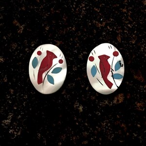 May include: A pair of oval-shaped earrings with a cardinal bird design. The birds are red, with blue leaves and red berries on a white background. The earrings are set against a dark, speckled surface.