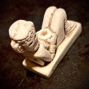 May include: A small, off-white, textured figurine of a reclining figure resting on a rectangular base. The figure has a rounded head with detailed markings, bent knees, and arms positioned across the torso, featuring a weathered, matte finish.