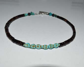 May include: A necklace featuring dark brown, flat, circular beads. The necklace has a series of turquoise-colored, patterned beads in the center. Small turquoise and black beads are interspersed throughout the necklace, which is secured with a silver clasp.