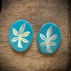 May include: Two oval-shaped turquoise earrings with a white leaf design. The leaf is inlaid with a mother-of-pearl material, creating a shimmering effect. The earrings are set against a wooden background.