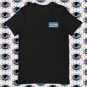 Könnte beinhalten: Schwarzes T-Shirt mit einem blauen Namensschild, auf dem "Frag mich nach meinem Trauma" steht.