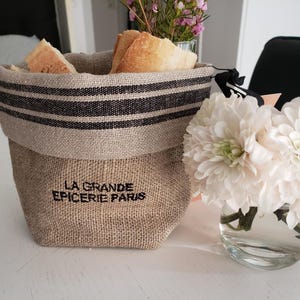 May include: A rustic burlap bread basket with black stripes and the text "LA GRANDE EPICERIE PARIS" holds slices of bread. A vase of white flowers sits nearby. The basket is on a white surface.