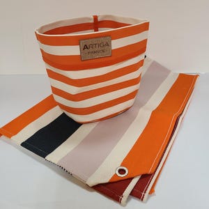 Puede incluir: Una cesta de tela a rayas naranjas y blancas, con una etiqueta "ARTIGA FRANCE", se encuentra sobre una tela a rayas a juego. La tela presenta rayas en naranja, blanco, negro, gris y burdeos.