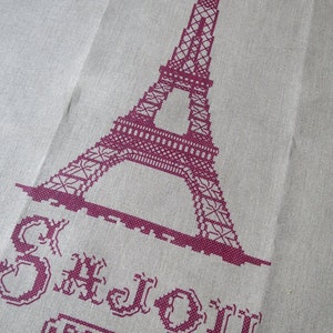 May include: Embroidered design of the Eiffel Tower in burgundy on a beige fabric background. The text "Je t'aime Paris" is also embroidered in burgundy below the tower.