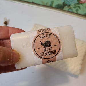 Può includere: Una saponetta bianca con un'etichetta marrone e bianca che recita "SAVON MUCUS D'ESCARGOT" con un'illustrazione di una lumaca. Il sapone è rettangolare e sembra non profumato. Sullo sfondo è visibile un piccolo asciugamano bianco e strutturato.