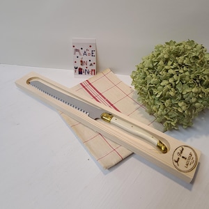 May include: A serrated cheese knife with a cream-colored handle and gold accents, presented in a wooden box. The knife rests on a red and white checkered cloth. A card with "MADE IN FRANCE" is visible.