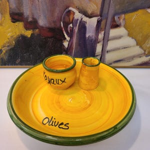 May include: A yellow ceramic serving set featuring a large round bowl with a green rim and the word Olives painted in black. Inside the bowl sits a small yellow pot with a green rim labeled Joyaux and a plain yellow cylindrical container.
