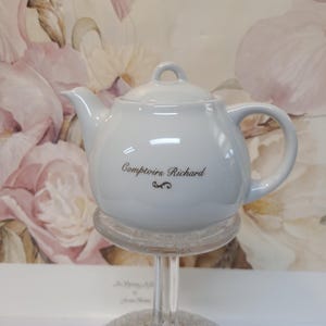 Könnte beinhalten: Eine weiße Keramik-Teekanne mit Deckel, Ausguss und Henkel. Auf der Teekanne steht in schwarzer Schrift "Comptoirs Richard". Die Teekanne steht auf einem transparenten Glasständer.