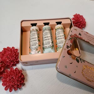 Könnte beinhalten: Eine rosa Metalldose mit drei blumig duftenden Handcremes. Die Dose hat ein Blumenmuster und den Text "Collection Hand Cream" an der Seite. Die Handcremes sind mit verschiedenen Blumendüften gekennzeichnet: "Geranienrose", "Kostbarer Jasmin" und "Orangenblüte".