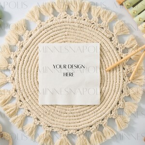 May include: A white card with the text "Your Design Here" on a round, beige, macrame placemat with tassels. The placemat is surrounded by various baby toys, including a wooden xylophone, a green beaded necklace, and a crocheted bunny.