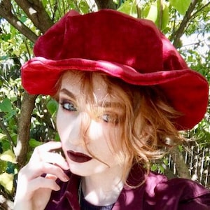 May include: A woman with red hair wearing a red velvet hat. The hat has a wide brim and a rounded crown.