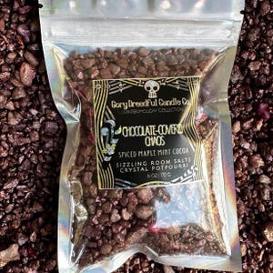 May include: A bag of chocolate-covered rock candy potpourri with a skull logo and the text "Gory Dreadful Candle Co. Winter/Holiday Collection Chocolate-Covered Chaos Spiced Maple Mint Cocoa Sizzling Room Salts Crystal Potpourri 6 oz 170 g".